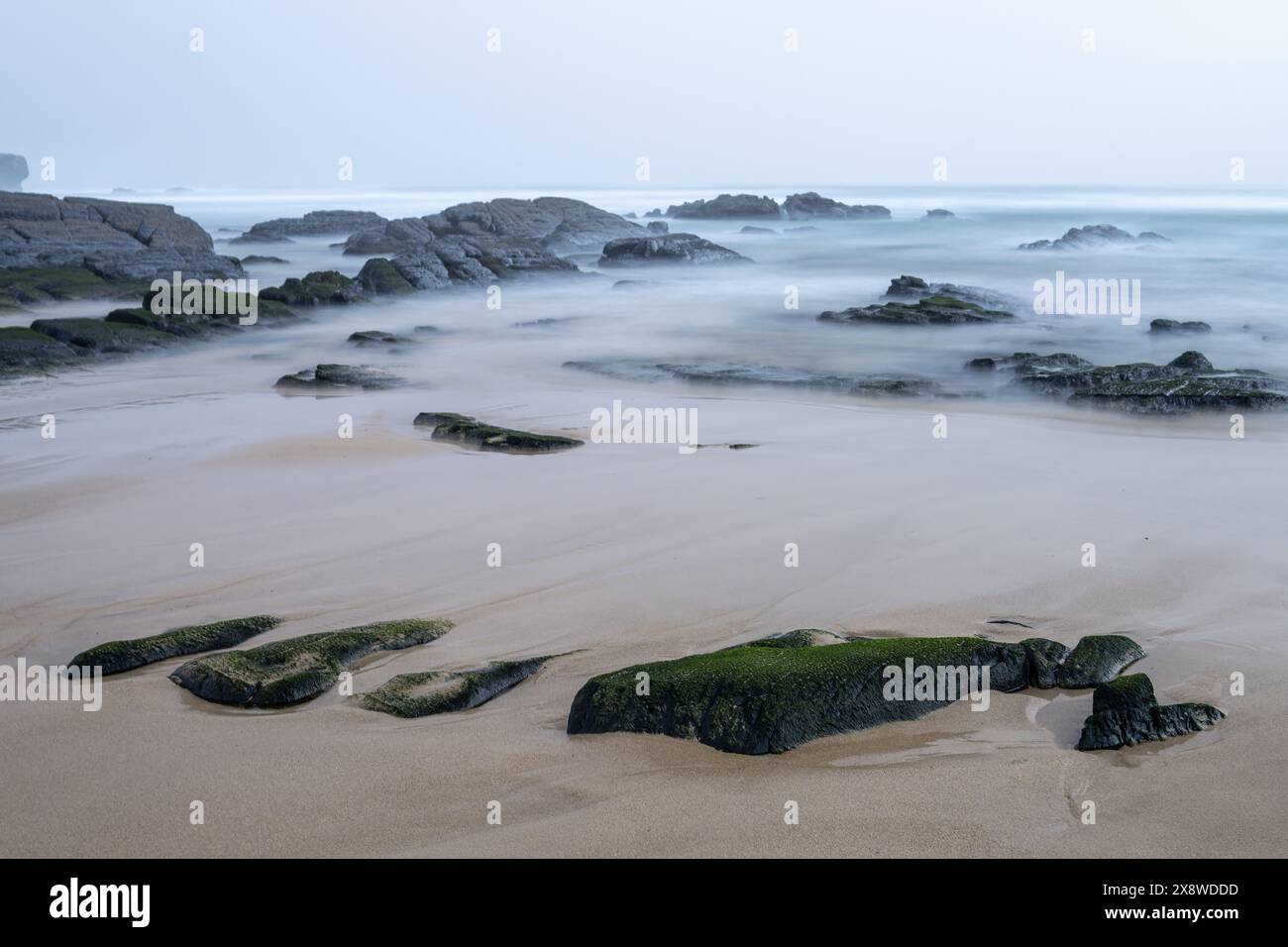 Eine ruhige Szene am Amoreira Beach in Portugal, mit nebelbedeckten Felsen und sanften Wellen, die sanft gegen die Sandküste schlagen. Die ruhige Atmosphäre Stockfoto