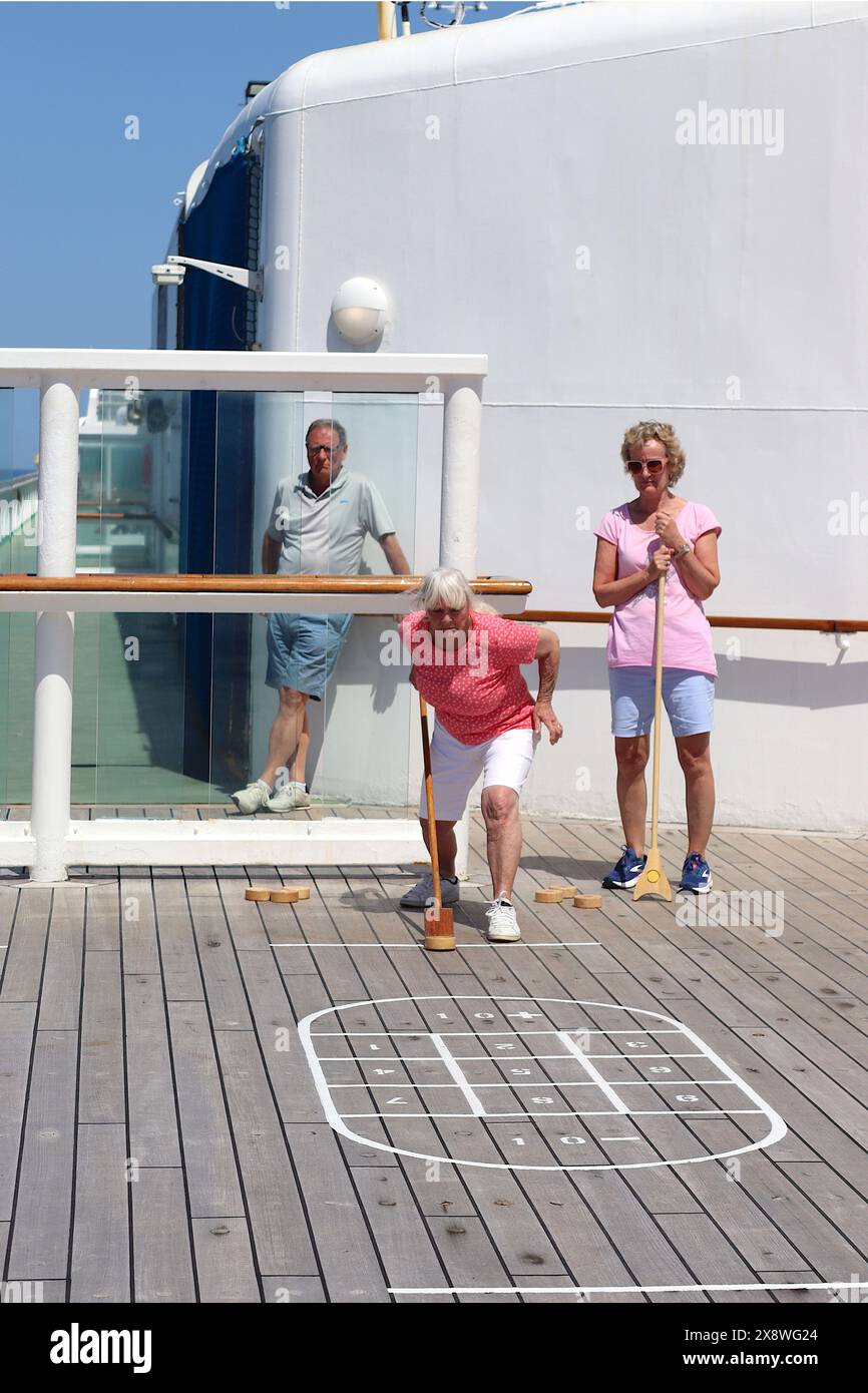 Eine Dame bereitet sich darauf vor, einen hölzernen Puck auf dem Shuffleboard-Platz in Richtung des Punktbereichs zu starten, während ihr Gegner und ein männlicher Kreuzfahrtgast auf dem Spielfeld zuschauen. Stockfoto