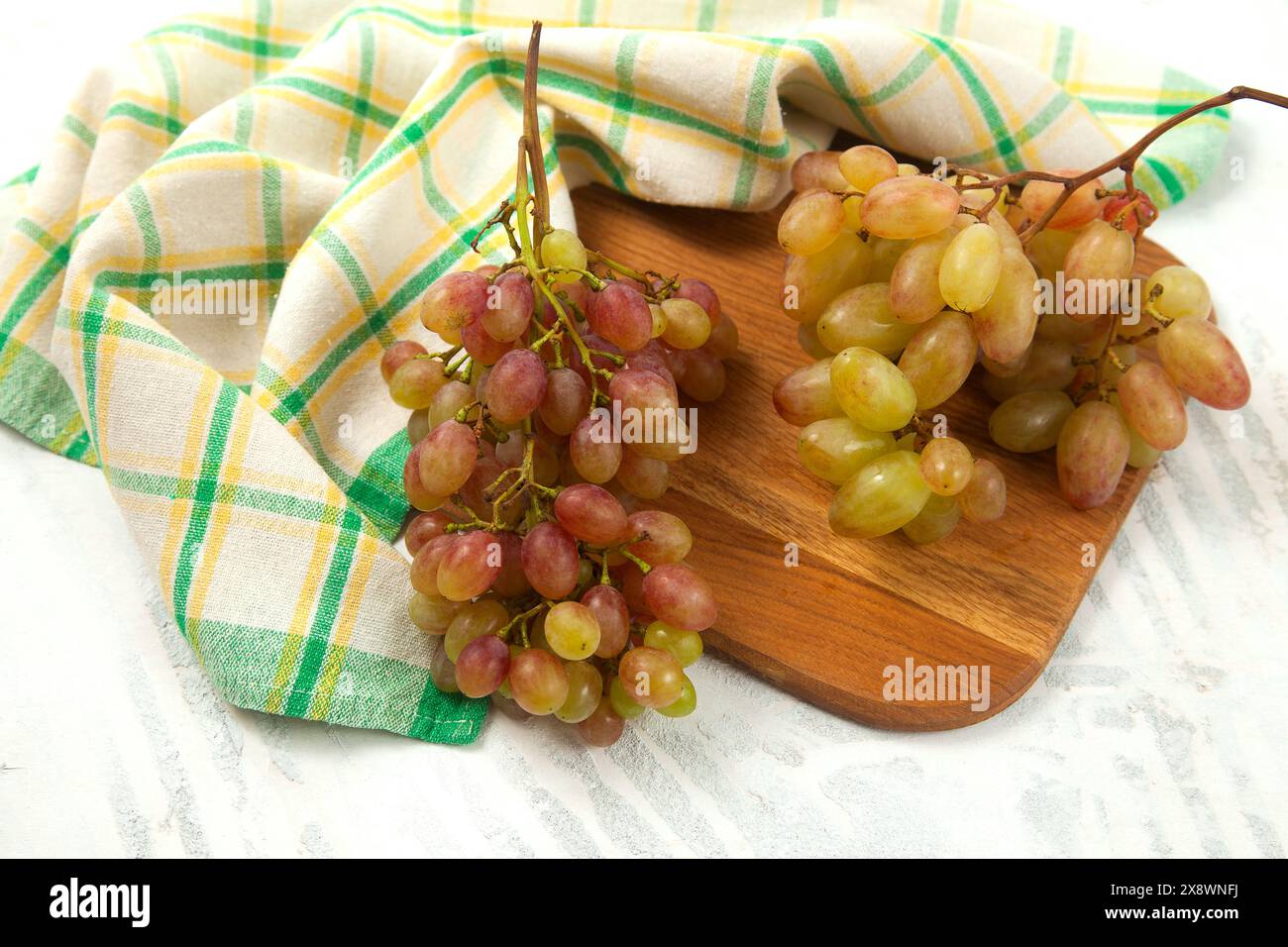 Schneidebrett mit einem großen Haufen reifer rosafarbener Trauben auf weiß. Rosafarbene Weintrauben mit blauem Küchentuch auf weißem hölzernem Hintergrund. Stockfoto