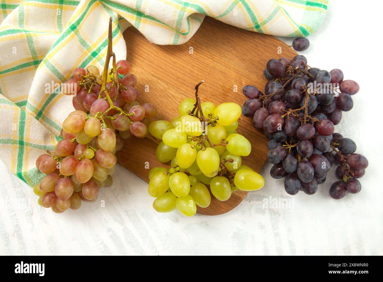 Schneidebrett mit einem großen Haufen reifer grüner, rosa und dunkelblauer Trauben auf weiß. Weintrauben mit grünem Küchentuch auf weißem hölzernem Hintergrund. Stockfoto