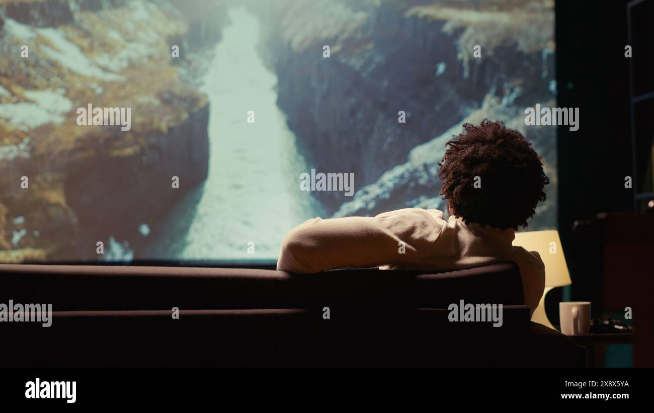afroamerikanischer Mann, der sich Naturdokumentationen mit atemberaubender Kinematographie auf einem riesigen Fernsehbildschirm ansieht. Person genießt Landschaftsaufnahmen auf Breitbildfernseher, entspannt auf der Couch Stockfoto