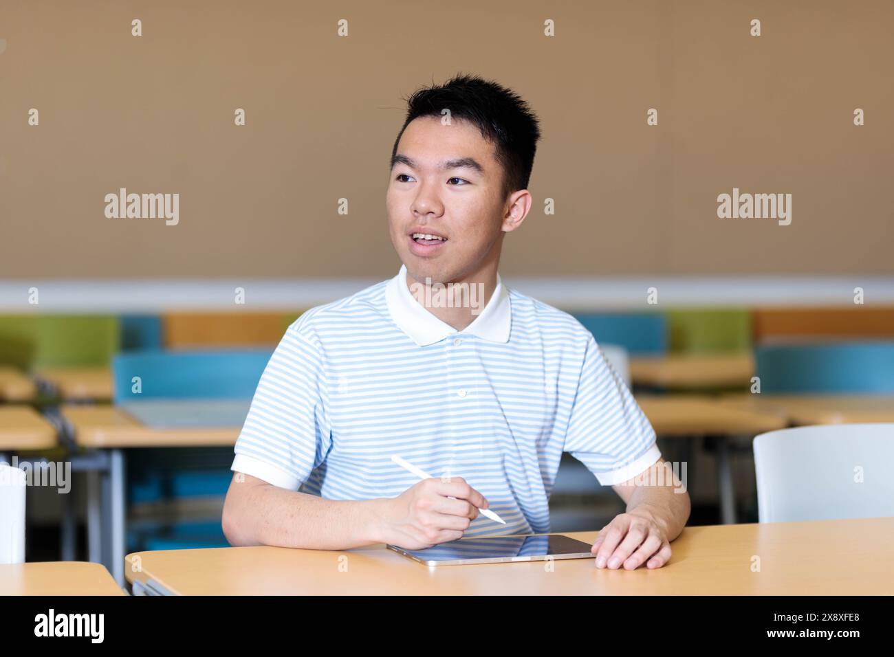 Ein junger Student im Teenageralter saß an einem Tisch im Klassenzimmer, lächelte und schaute sich mit seinem Tablet vor sich herum. Stockfoto