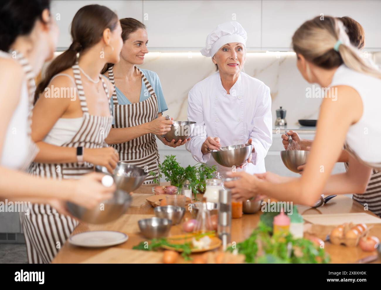 Ältere Chefkochin mit Schüssel in den Händen erzählt Frauen bei kulinarischen Kursen über die Traditionen der Küche Stockfoto