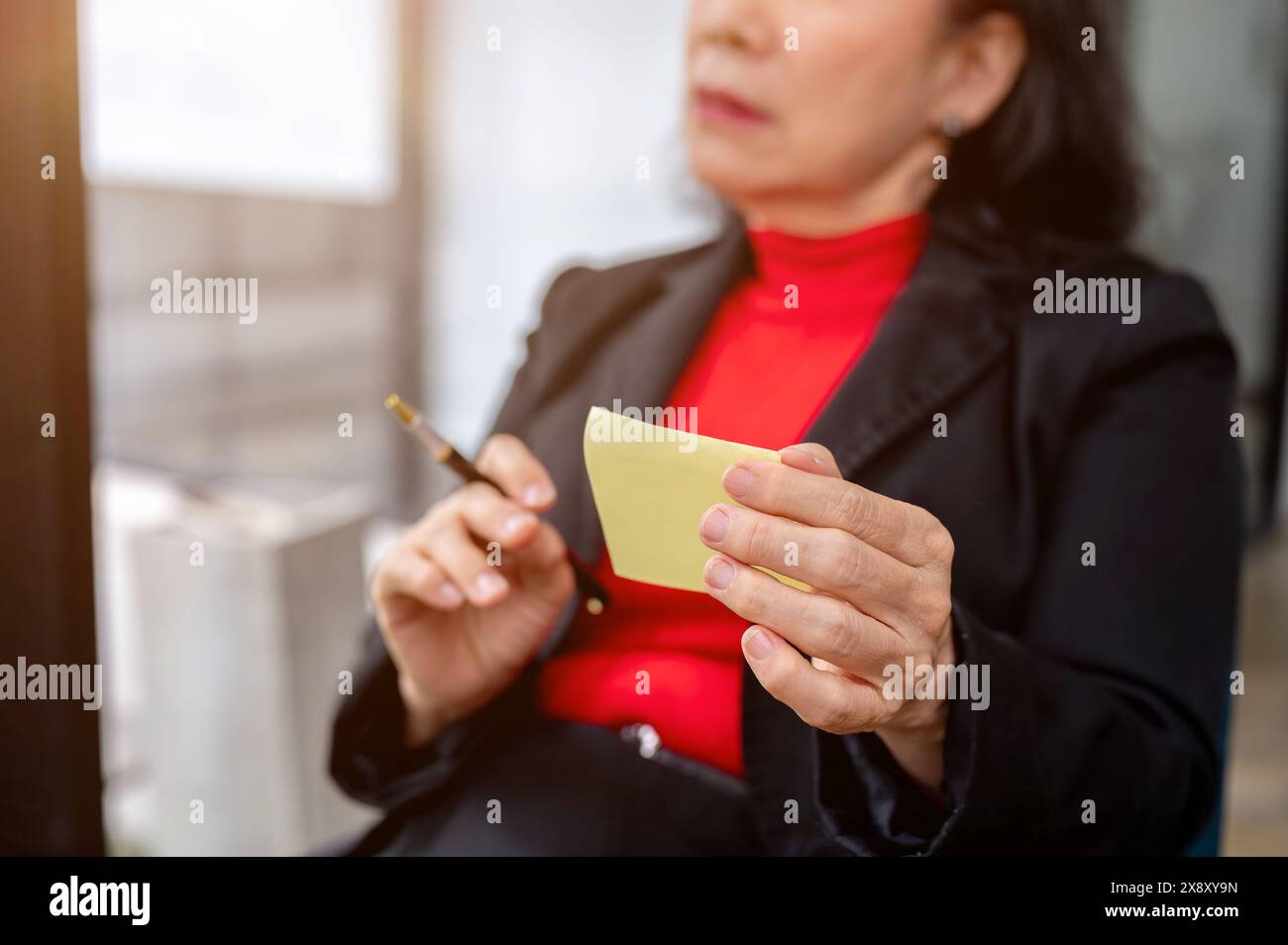 Ein selektives Bild einer nachdenklichen asiatischen Geschäftsfrau, die eine Haftnotiz und einen Stift in der Hand hält, die an ihrem Schreibtisch im Büro in Gedanken verloren ist. Stockfoto