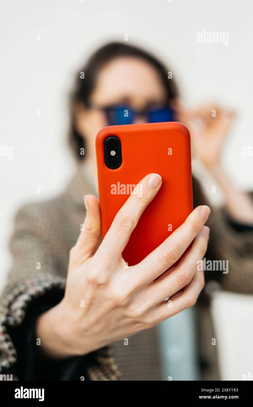 Frau mit Sonnenbrille und kariertem Mantel, die ein Selfie mit ihrem roten Smartphone macht und ihre Brille mit einer Hand anpasst. Stockfoto