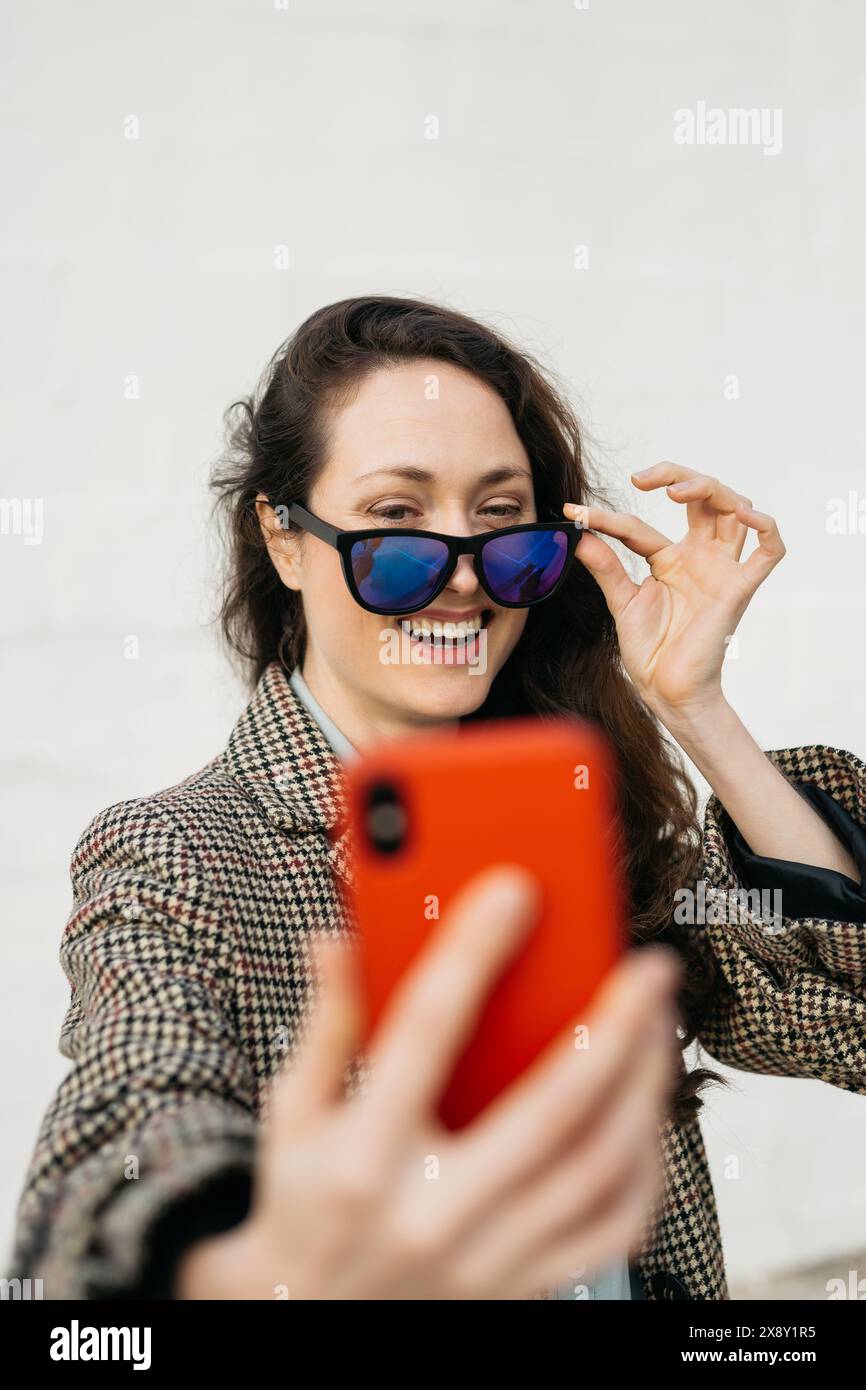 Eine Frau mit Sonnenbrille und kariertem Mantel macht ein Selfie mit ihrem roten Smartphone, lächelt und passt ihre Brille mit einer Hand an. Stockfoto