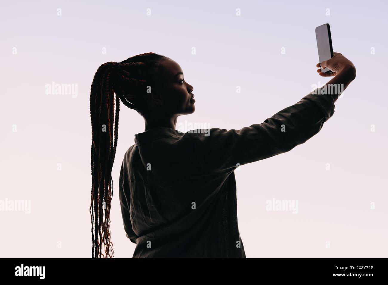 Eine selbstbewusste Frau aus der Generation Z macht ein Selfie mit einem Smartphone. Sie steht in einem Studio vor weißem Hintergrund und trägt einen Pferdeschwanz mit Brai Stockfoto
