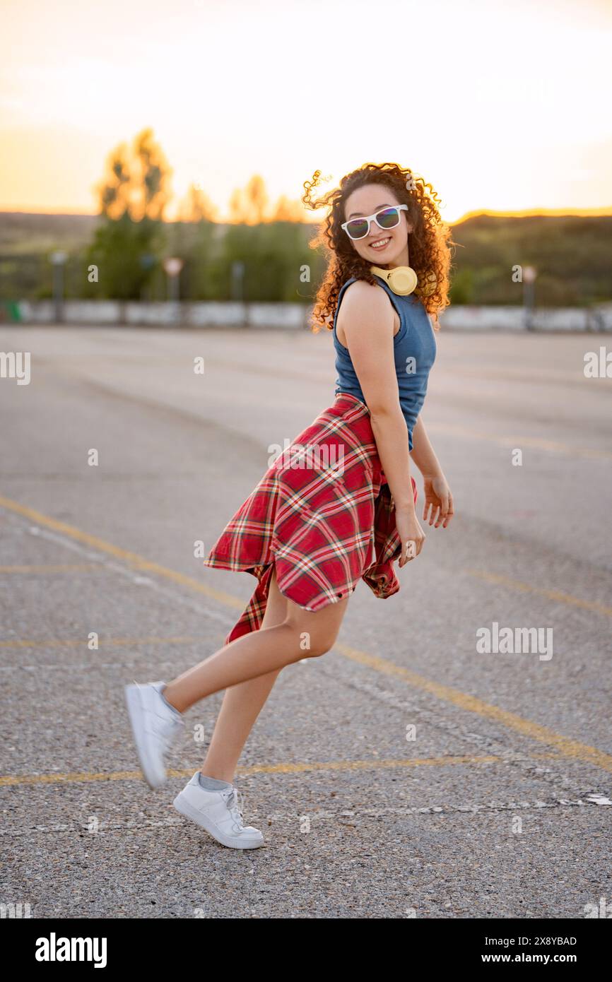 Fröhliche junge Frau in Sonnenbrille und Kopfhörern, die bei Sonnenuntergang auf einem Parkplatz spielerisch herumwirbelt. Stockfoto