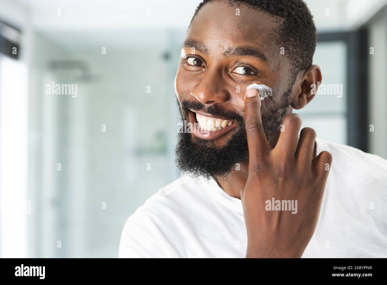 Zu Hause, Afroamerikaner, der Feuchtigkeitscreme aufträgt, lächelt in die Kamera. Mit Bart und kurzen Haaren trägt er ein weißes Hemd und strahlt entspannt aus Stockfoto