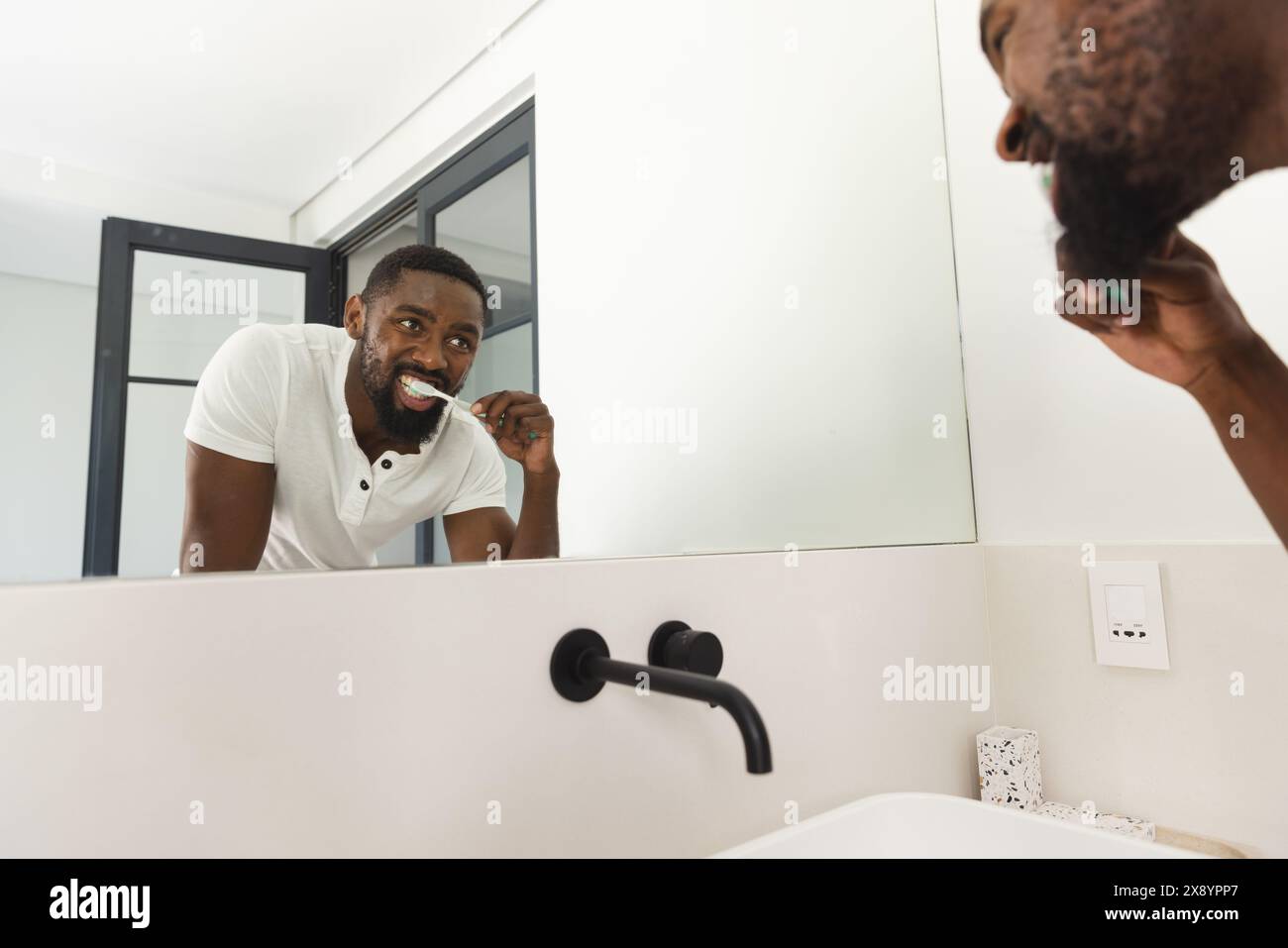 Zu Hause im Badezimmer, Afroamerikaner putzt Zähne. Mit lässigem weißem Hemd, gepflegtem Aussehen, Blick in den Spiegel, unverfälscht Stockfoto