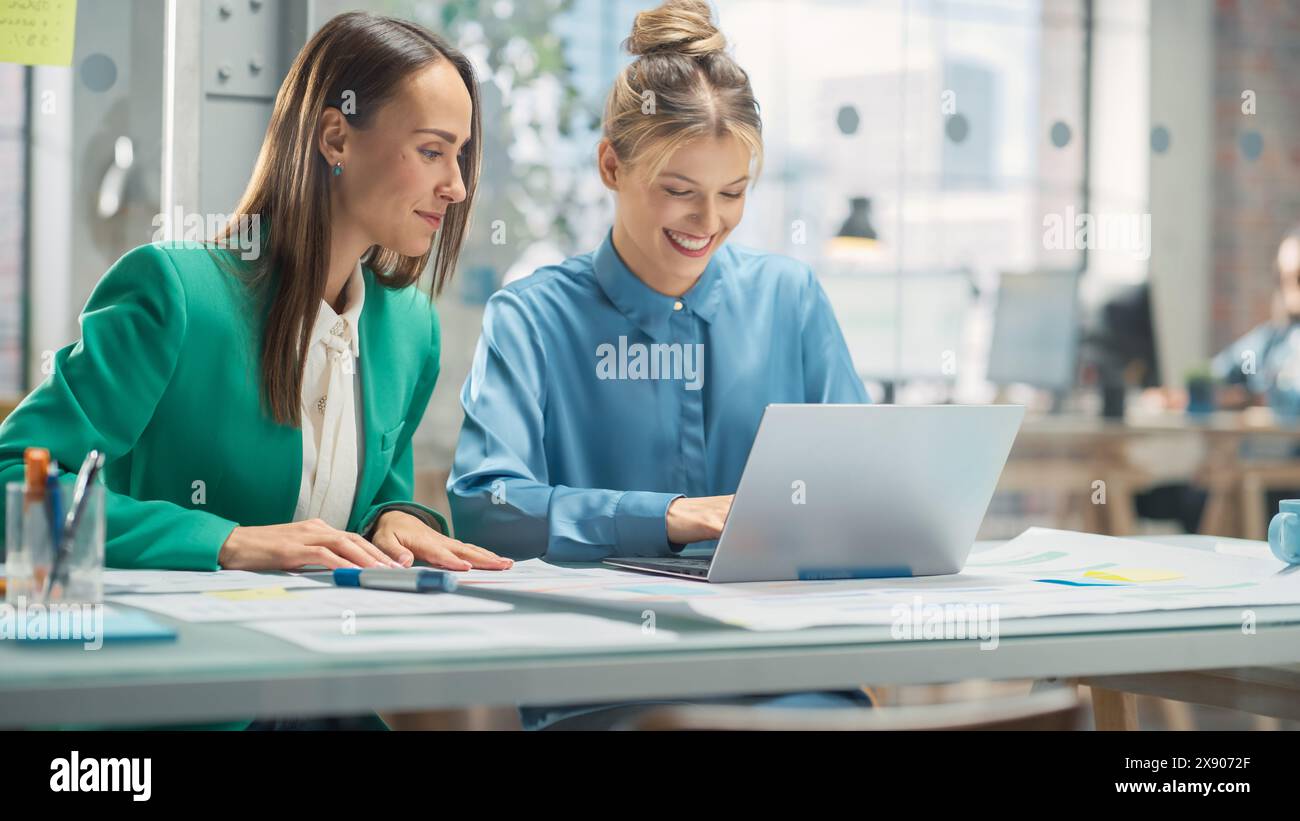 Porträt zweier kreativer Kollegen, die in Office Hub auf den Laptop-Bildschirm zeigen. Weiße Designerin bespricht die Arbeit mit der Assistentin und wählt die Marketingstrategie aus Stockfoto