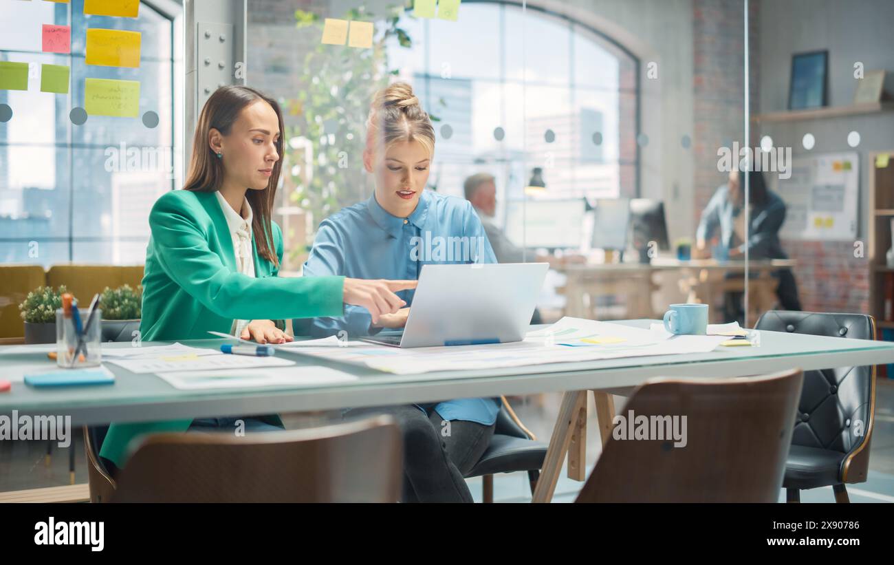 Porträt zweier kreativer Kollegen, die in Office Hub auf den Laptop-Bildschirm zeigen. Weiße Designerin bespricht die Arbeit mit der Assistentin und wählt die Marketingstrategie aus Stockfoto