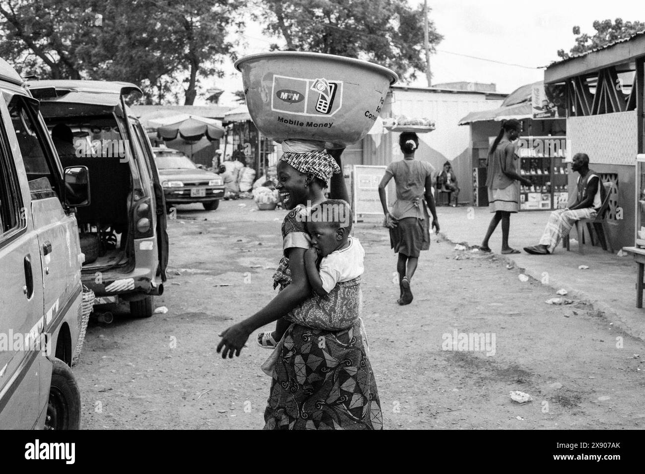 Kayayei Frau mit einer großen Schüssel auf dem Kopf, während sie ein Kind auf einem geschäftigen Markt in Accra, Ghana, hält Stockfoto