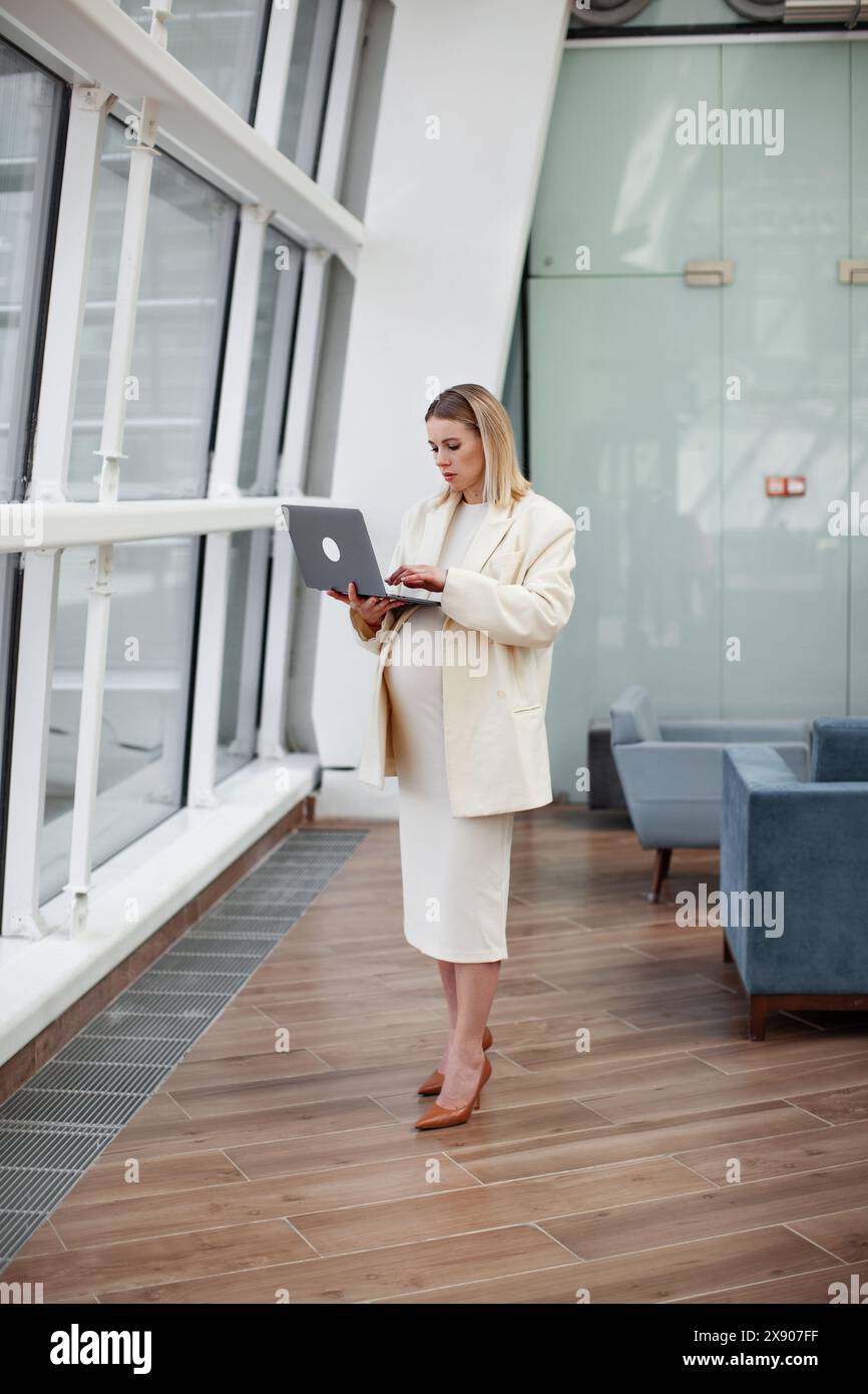 Schwangere Geschäftsfrau, die selbstbewusst an einem Laptop in einem modernen Business-Office mit hellblauem Hintergrund arbeitet, einen stilvollen Anzug und Blazer trägt, re Stockfoto