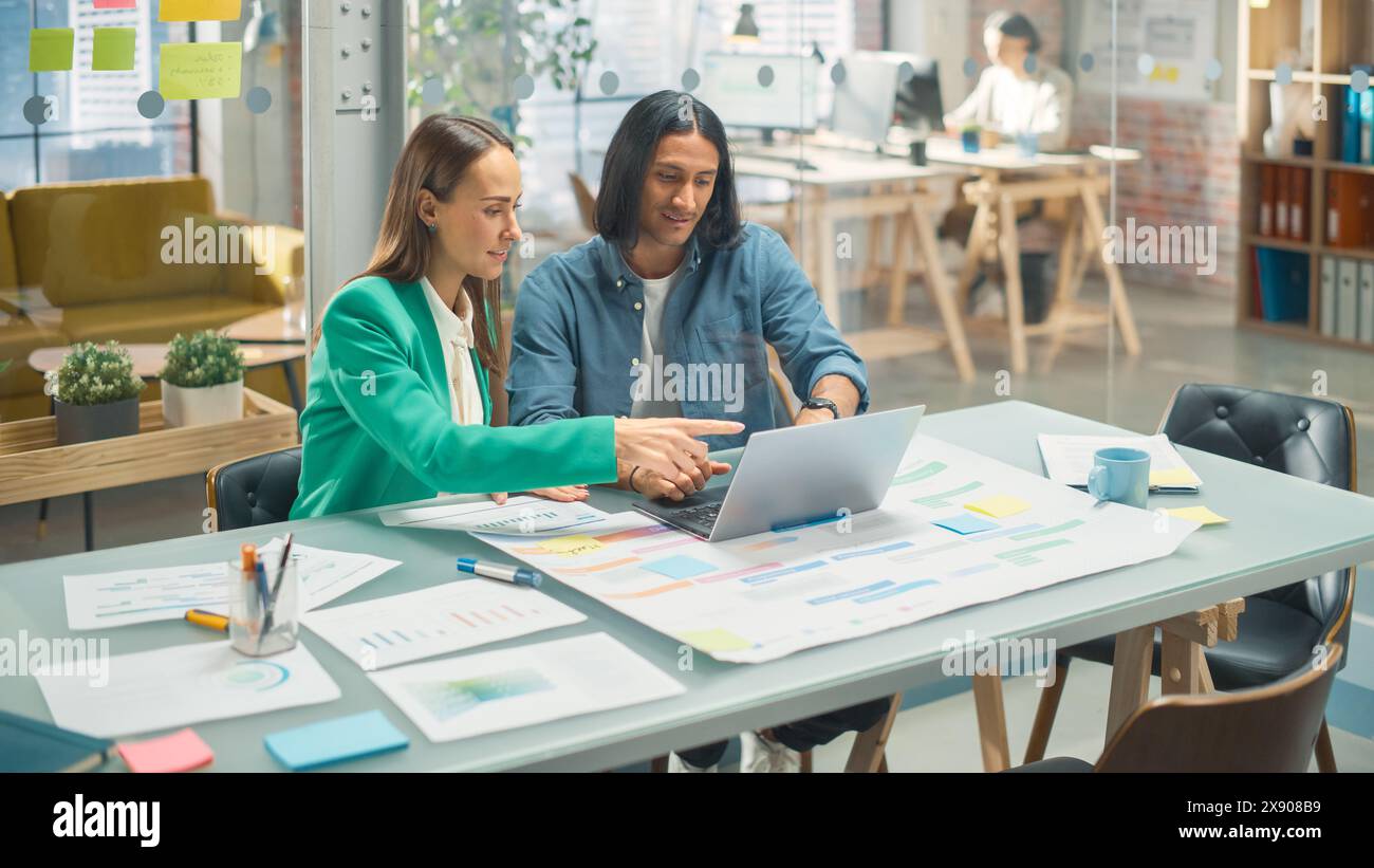 Porträt zweier kreativer Kollegen, die in Office Hub auf den Laptop-Bildschirm zeigen. Weiße Designerin bespricht die Arbeit mit der Assistentin und wählt die Marketingstrategie aus Stockfoto