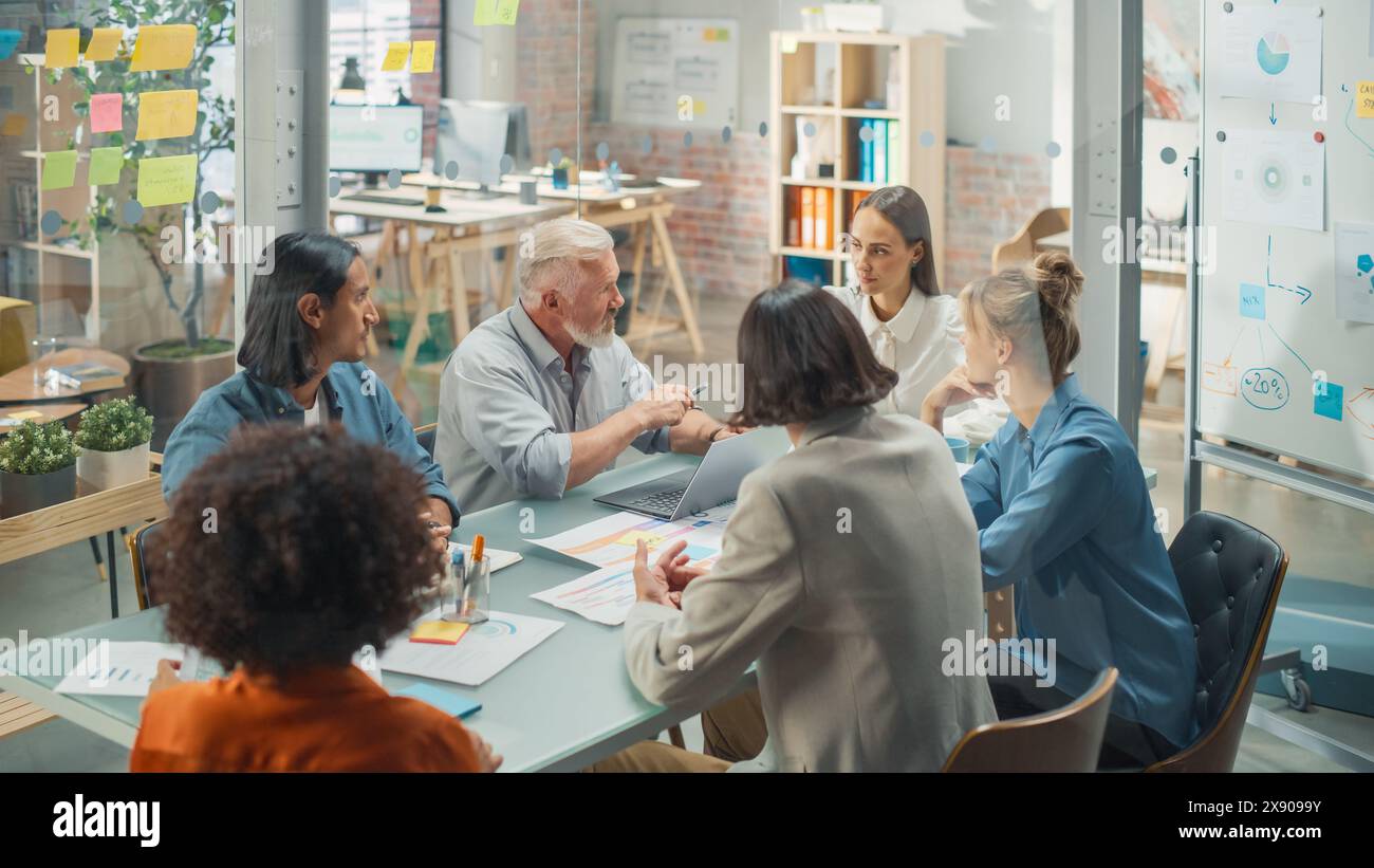 Im Konferenzraum diskutieren Unternehmer und Spezialisten am Konferenztisch über Statistiken und Diagramme, lösen Probleme unter Leitung des Senior Lead. Sie verwenden Laptop Stockfoto