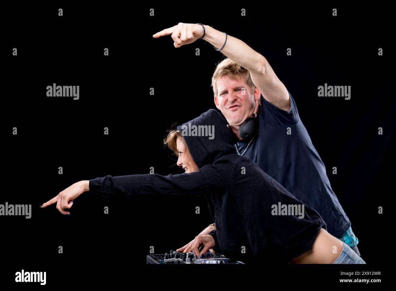 DJ-Paar, lässig gekleidet, genießt eine spielerische Interaktion während der Arbeit am Mixer. Die Frau in einem Kapuzenpulli lächelt breit, und der Mann gibt Enthusi Stockfoto