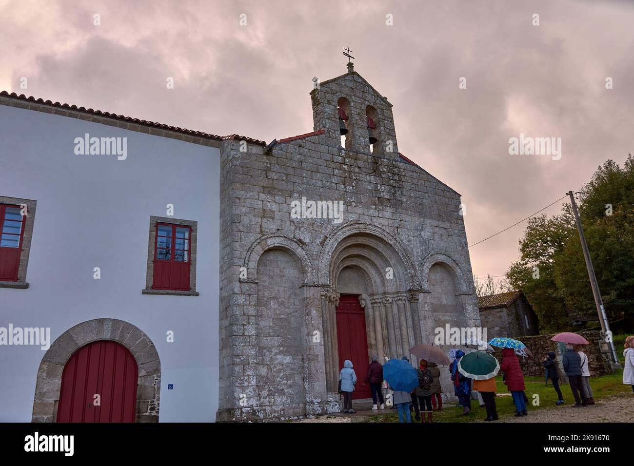 Mit Sonnenschirmen besichtigen Sie eine historische Steinkirche mit roten Türen unter einem bewölkten Himmel, die kulturelle Interessen und Kulturerbe widerspiegelt Stockfoto