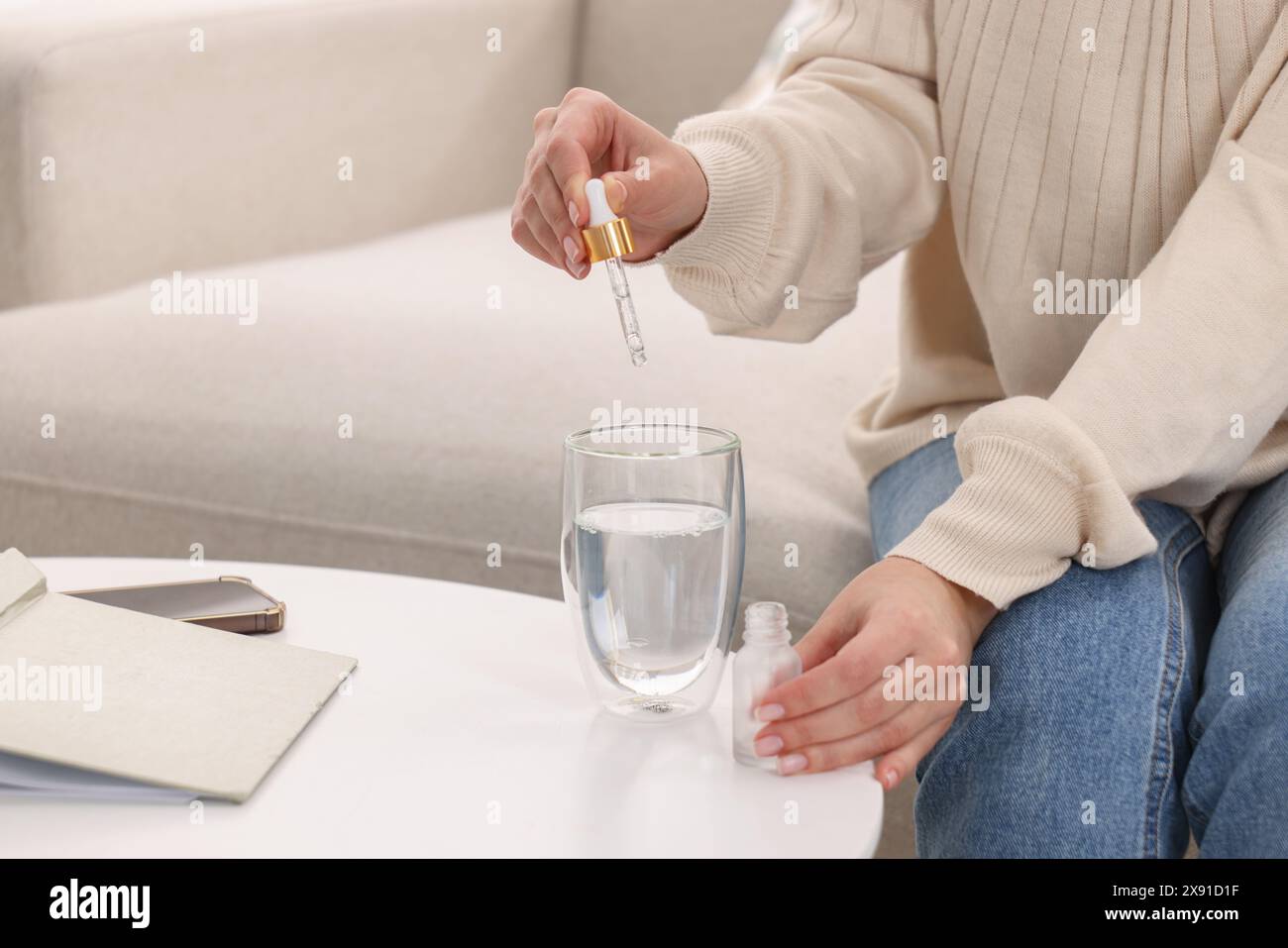 Frau tropft Nahrungsergänzungsmittel in ein Glas Wasser auf einem weißen Tisch drinnen, Nahaufnahme Stockfoto