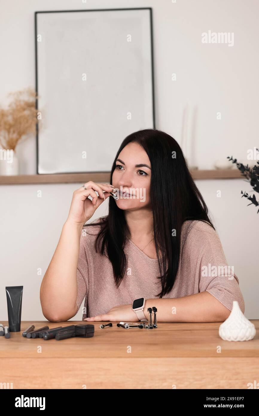 Eine Frau mit schwarzen Haaren und einem rosa Hemd sitzt an einem Tisch, umgeben von Make-up-Tools, und sieht durchdacht aus mit einem minimalistischen Dekor im Hintergrund Stockfoto