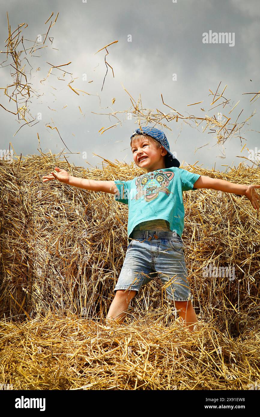 Ein fröhliches Kind, das auf Heuhaufen steht und die Arme gestreckt hat, ein blaues Hemd und Jeansshorts trägt, unter bewölktem Himmel, Weißrussland, Minsk Stockfoto