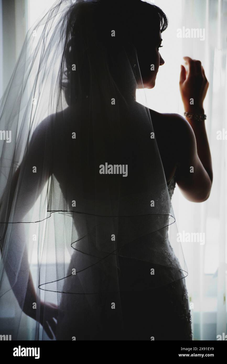 Silhouette einer Braut in einem Hochzeitskleid mit Schleier, die an einem sonnendurchfluteten Fenster steht und Eleganz und Vorfreude ausstrahlt Stockfoto