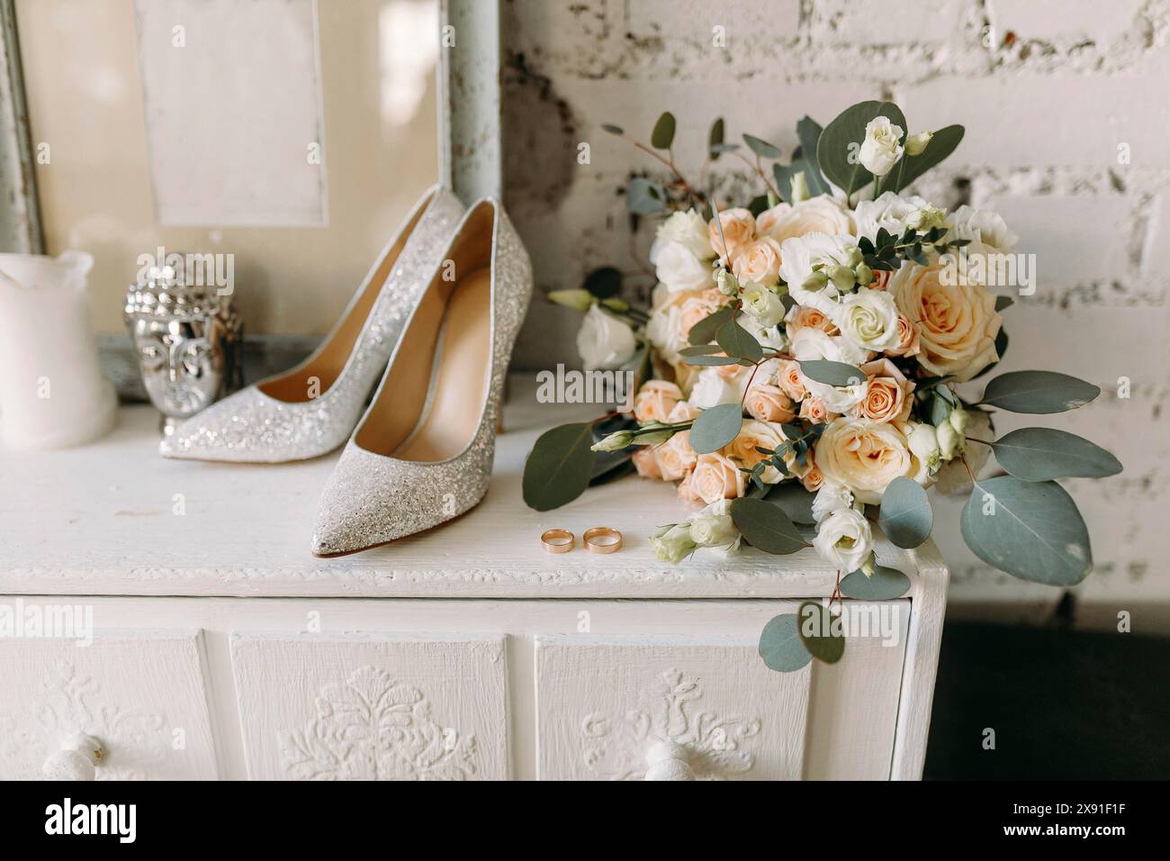 Elegante Hochzeitseinrichtung mit glitzernden Schuhen, Blumenstrauß und Ringen auf einer rustikalen weißen Kommode Stockfoto