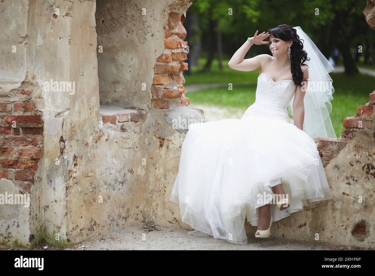 Eine Braut in einem weißen Hochzeitskleid mit Schleier, die draußen an einer alten Ziegelwand sitzt und unter natürlichem Licht lächelt. Weißrussland, Minsk Stockfoto