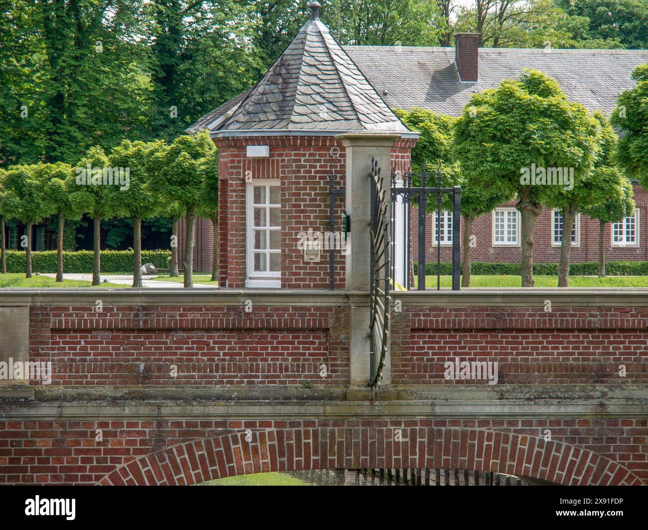 Ein kleines Backsteinhaus mit Tor, umgeben von Bäumen und einem gepflegten Innenhof an einem Sommertag, historisches Schloss mit grünem Park mit Stockfoto