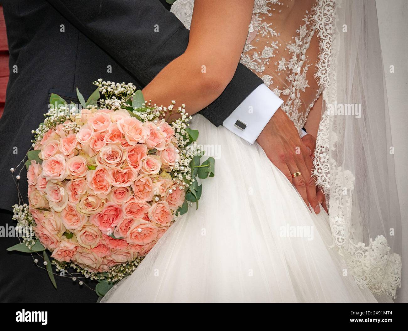 Eine Braut und ein Bräutigam umarmen ihre Liebe und ihr Engagement mit einem atemberaubenden Blumenstrauß aus rosa Rosen, einem zarten Spitzenkleid und einem detaillierten Anzug in der Sonne Stockfoto