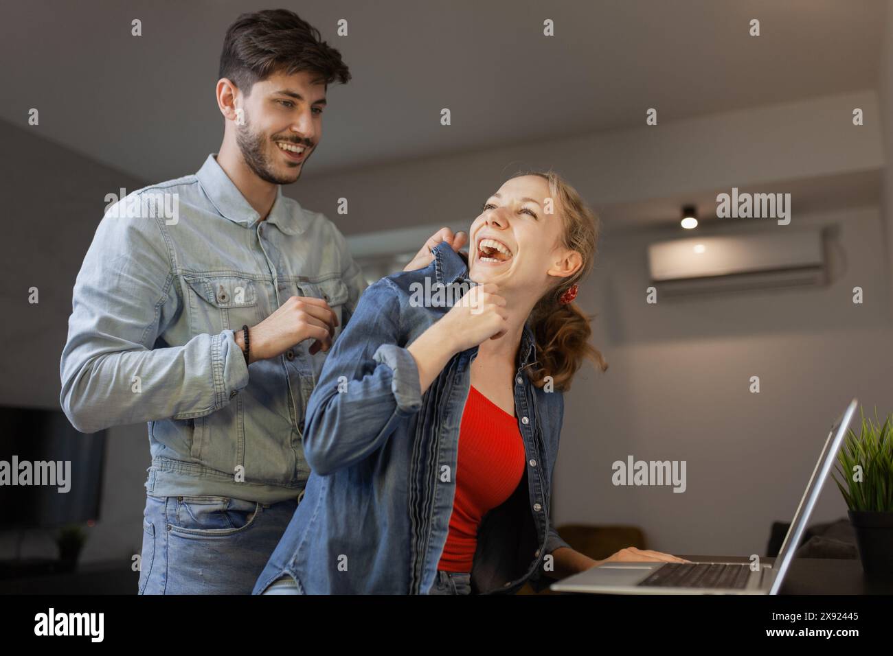 Ein junger Mann und eine junge Frau teilen eine fröhliche, spielerische Interaktion zu Hause mit einem Laptop im Hintergrund, der Glück und Verbindung ausstrahlt. Stockfoto