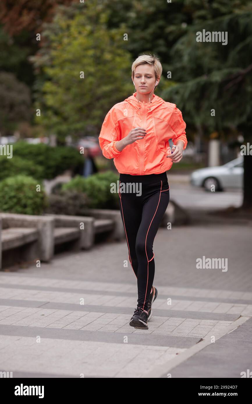 Die junge Joggerin in leuchtend orangefarbener Sportjacke, die in einem Stadtpark läuft, zeigt aktiven Lifestyle und Fitness-Routine. Stockfoto