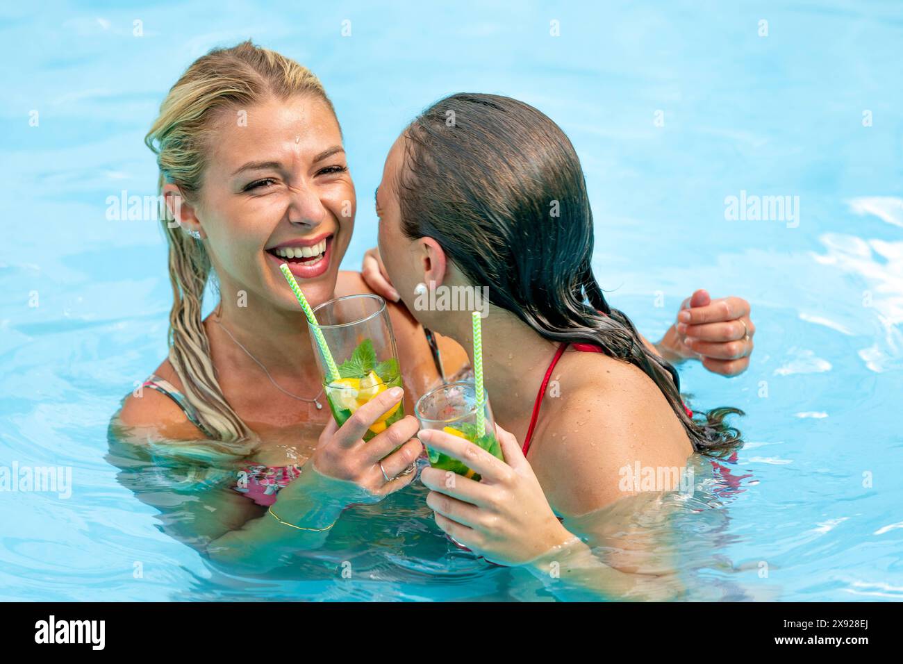 Zwei Freundinnen, die im Swimmingpool hekelten, während sie ein durstlöschendes Getränk sehen. Entspannung und Mittäterschaft am Pool mit Freunden. 016644 141 Stockfoto