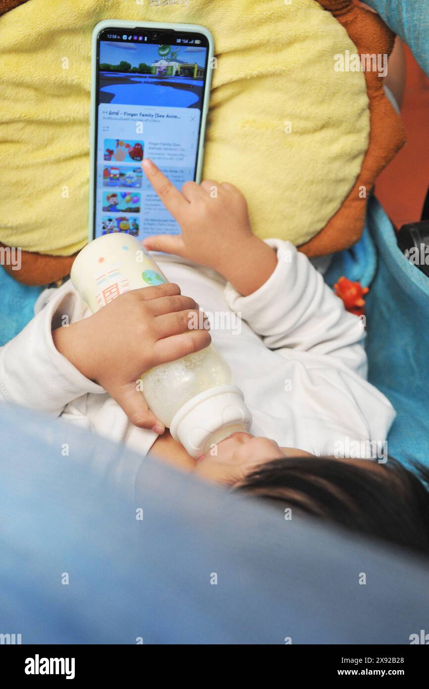 Kleines Kind in der Wiege mit seiner Flasche vor einem Telefonbildschirm. Baby und iphone 016812 005 Stockfoto