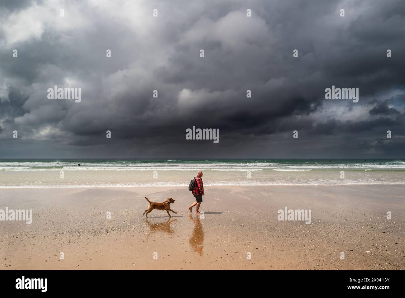 Ein Hundeführer und sein Hund laufen entlang des Fistral Beach, während sich dunkle, brütende Regenwolken der Küste von Newquay in Cornwall nähern. Stockfoto