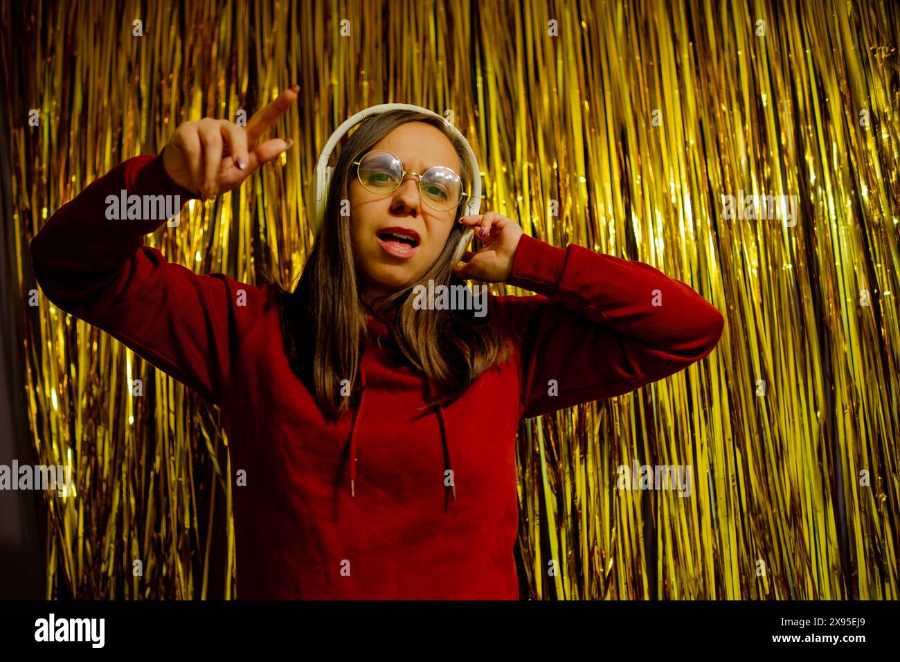 Eine Frau, die Brille und Kopfhörer trägt, tanzt energisch vor einem glänzenden goldenen Hintergrund Stockfoto