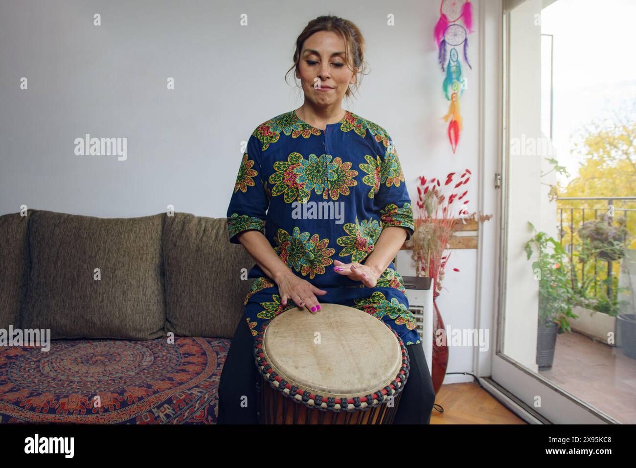 Vorderansicht einer erwachsenen Frau, die traditionelle afrikanische Musik zu Hause mit einer Djembe-Trommel am Balkon im Apartment praktiziert. Sie trägt AF Stockfoto