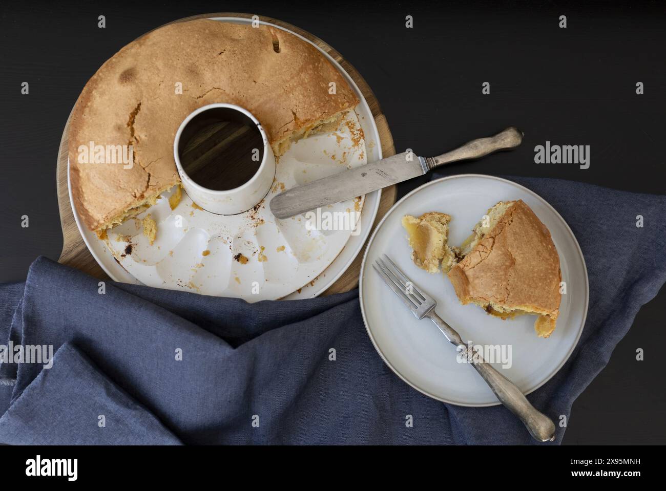 Blick von oben auf einen Apfelschwammkuchen in einer weißen abnehmbaren Dose, neben einem Stück Schwammkuchen auf einem hellen Teller mit Vintage-Messer und Gabel, auf einem Stockfoto