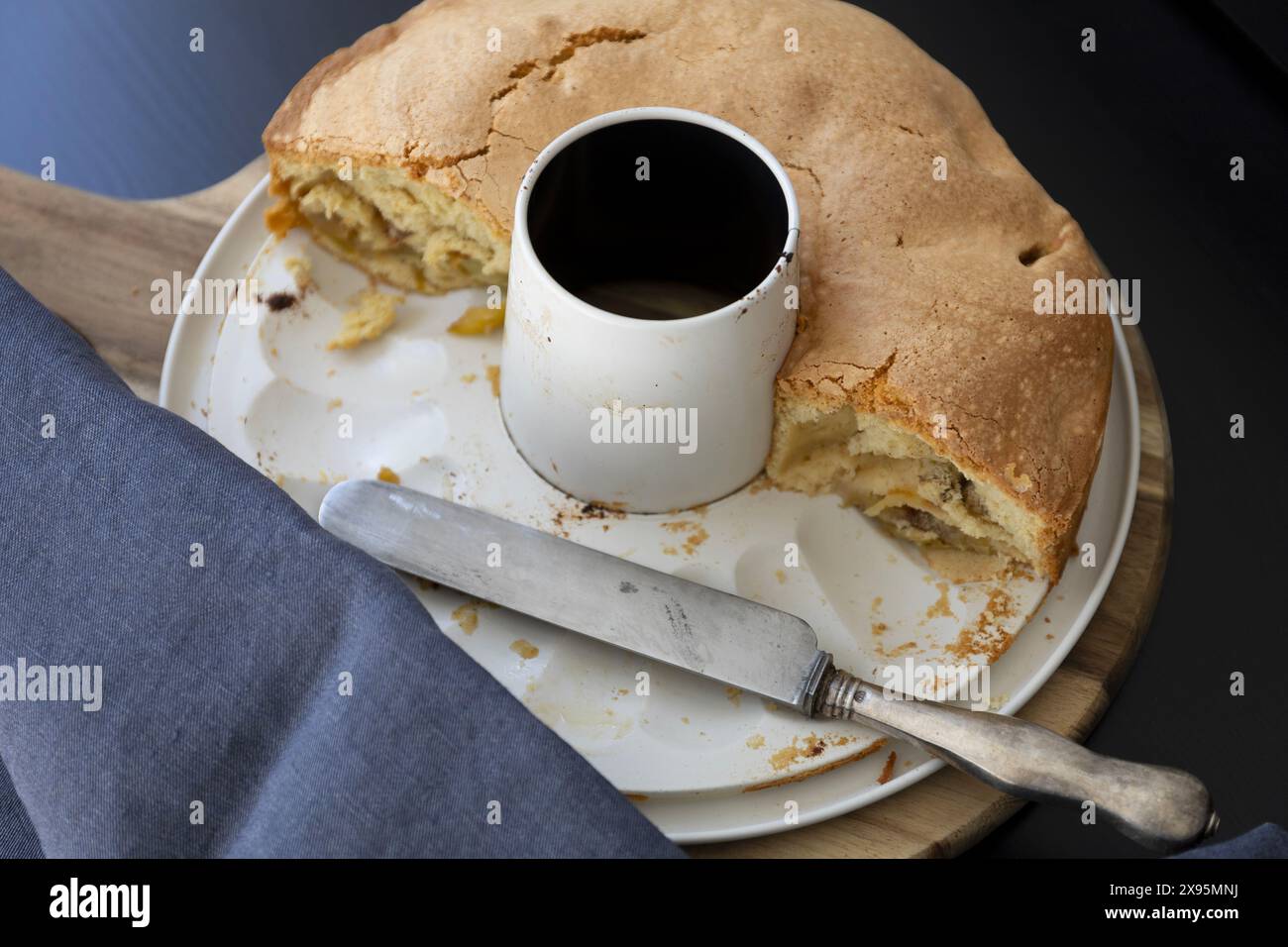 Blick von oben auf einen Apfelschwammkuchen in einer weißen abnehmbaren Dose mit Vintage-Messer auf schwarzem Hintergrund mit dunkelblauen Vorhängen in natürlichem Licht Stockfoto