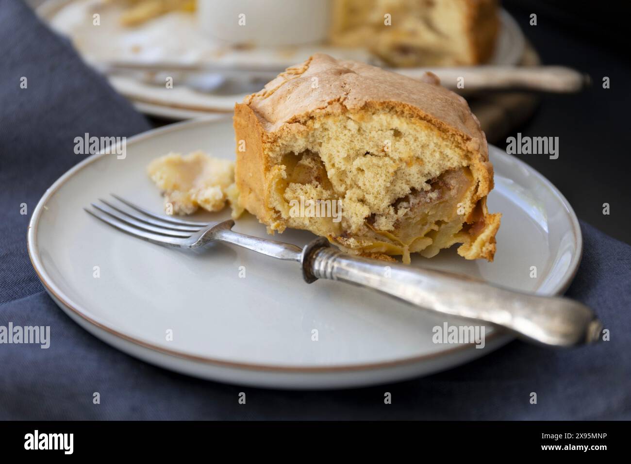 Ein Stück Biskuitkuchen auf einem hellen Teller mit Vintage-Gabel auf schwarzem Hintergrund mit dunkelblauen Vorhängen in natürlichem Licht Stockfoto