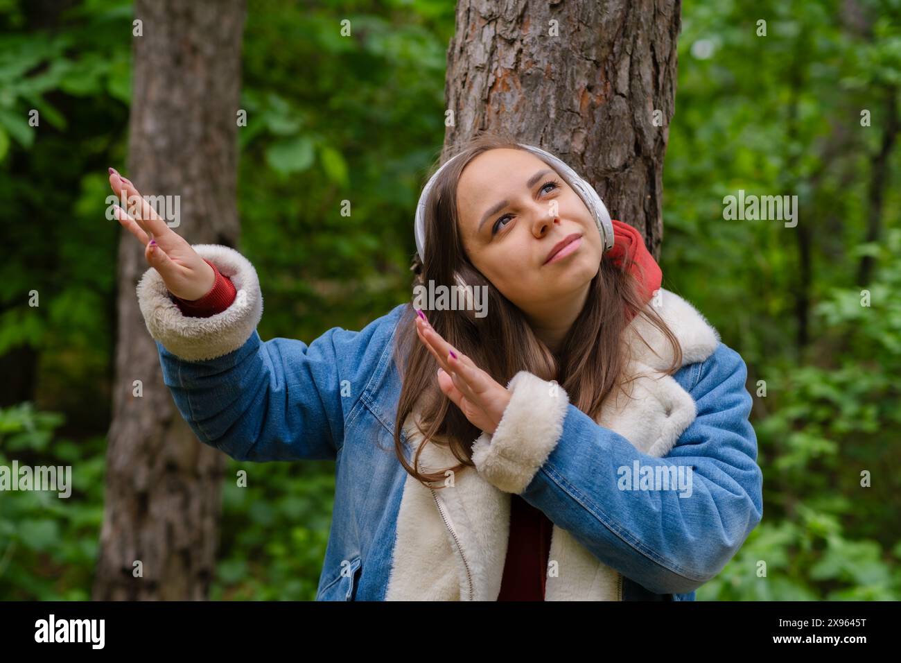Eine junge Frau lehnt sich an einen Baum, trägt Kopfhörer und genießt Musik im Wald an einem sonnigen Nachmittag Stockfoto
