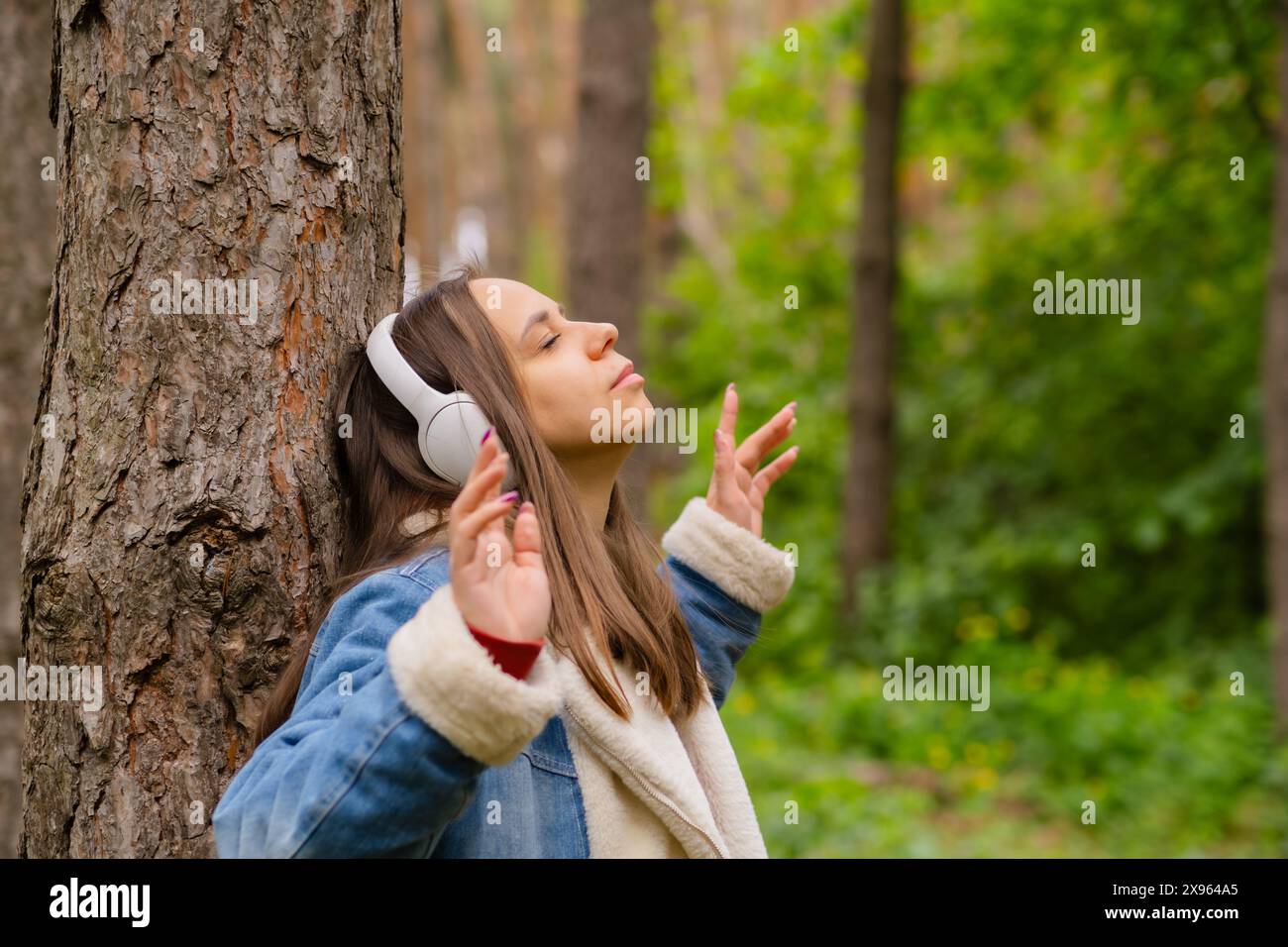 Eine junge Frau lehnt sich an einen Baum, trägt Kopfhörer und genießt Musik im Wald an einem sonnigen Nachmittag Stockfoto