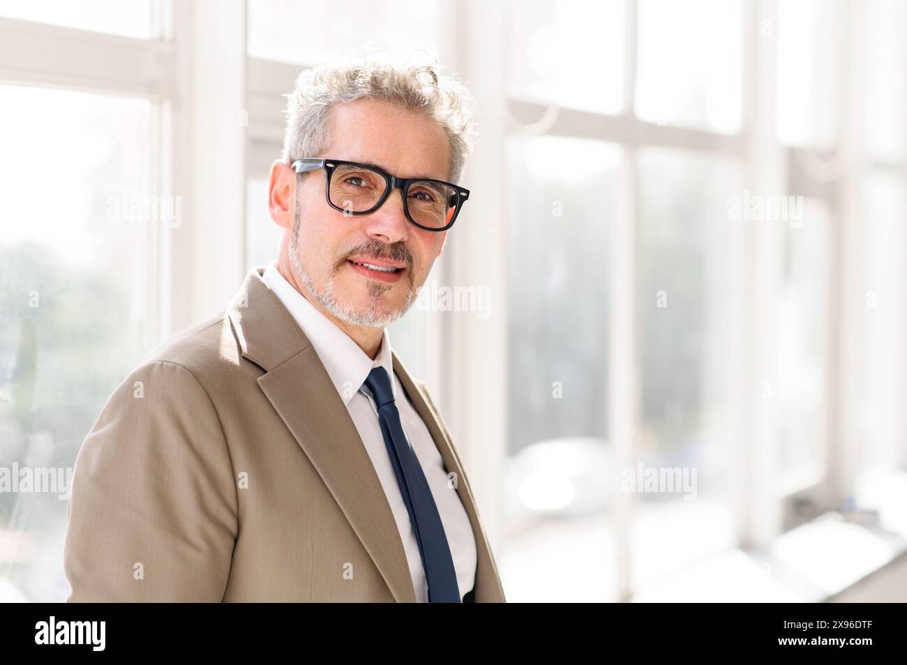 Ein geschickter, reifer Geschäftsmann mit grauen Haaren und einem warmen Lächeln steht selbstbewusst in einem gut beleuchteten Büro, sein scharfer Anzug und seine Brille sprechen von Weisheit und moderner Professionalität. Stockfoto