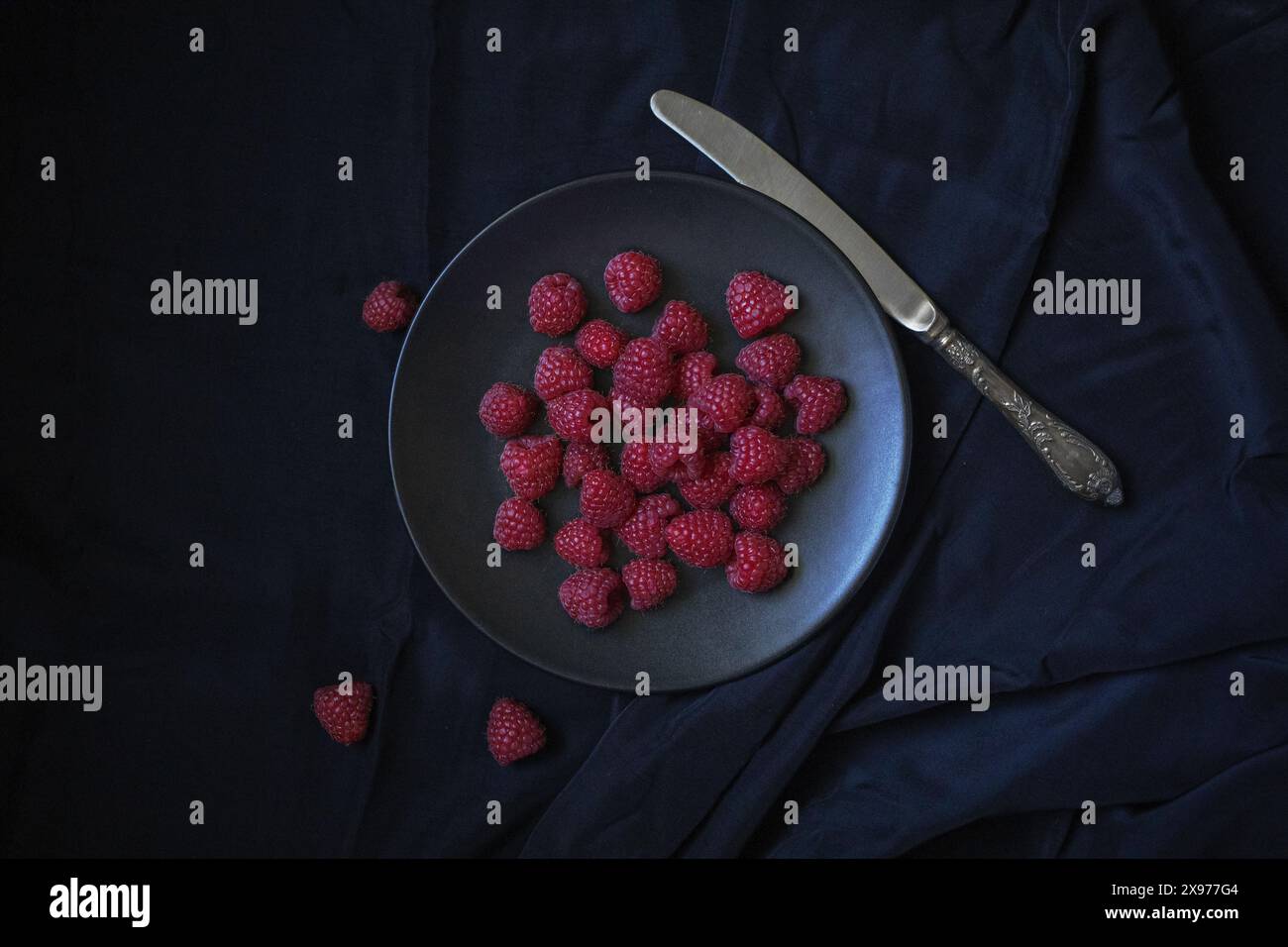 Einfache Tischfotografie von Himbeerbeeren auf einer schwarzen Keramikplatte mit Vintage-Messer auf einem schwarzen Seidenstoff Hintergrund Stockfoto