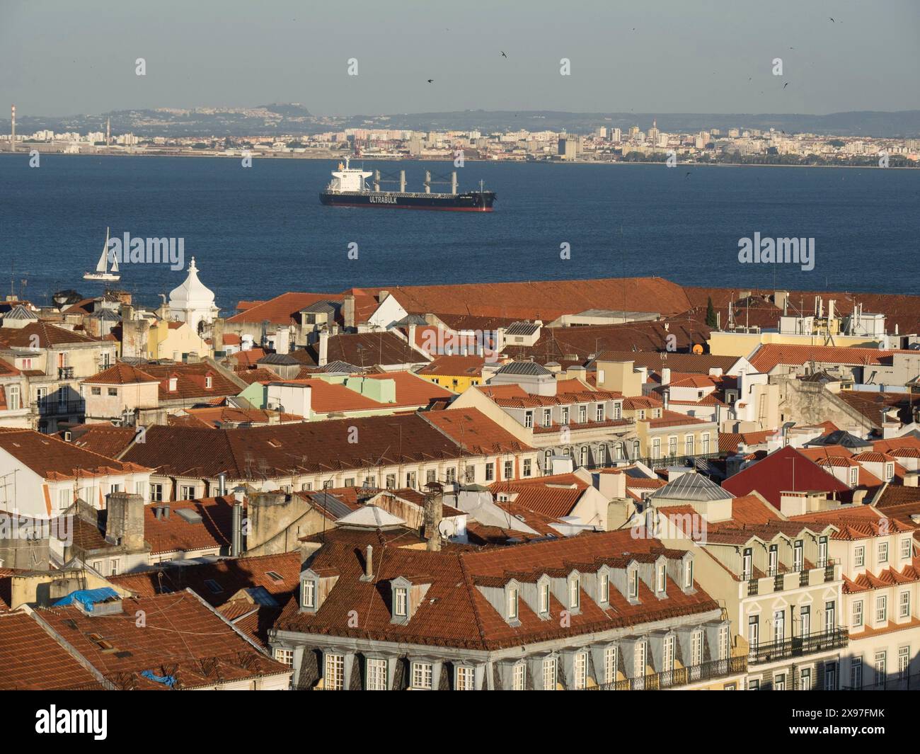 Weitwinkelblick auf eine historische Stadt mit roten Dächern und einem Frachtschiff im Hintergrund im Wasser, Blick auf die Altstadt von Lissabon am Fluss mit Stockfoto