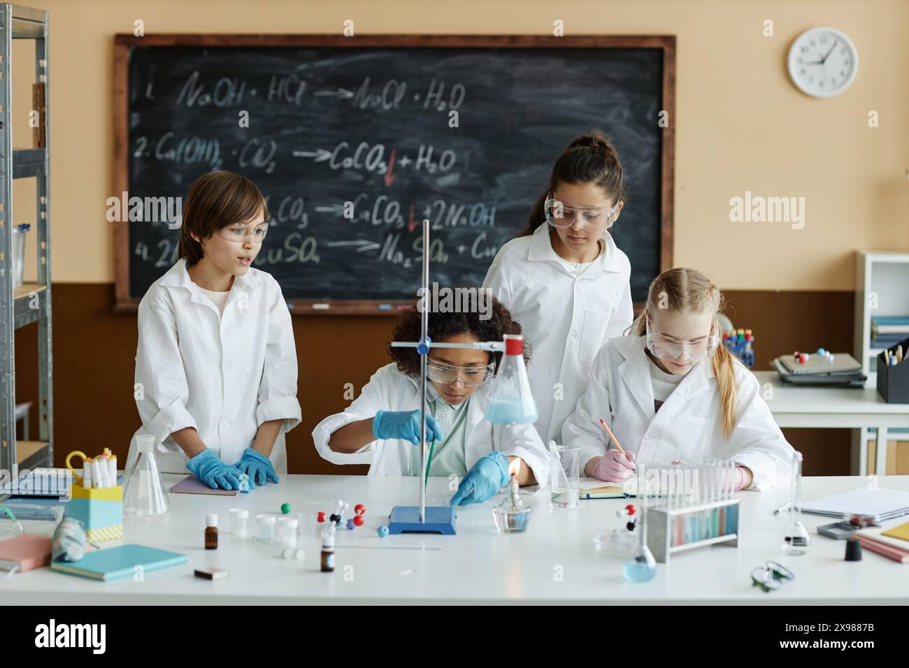 Gruppe von Mittelschüler mit Laborkittel und Schutzbrille, die im Unterricht am Tisch sitzen und stehen, um Experimente im Chemiekurs zu machen Stockfoto