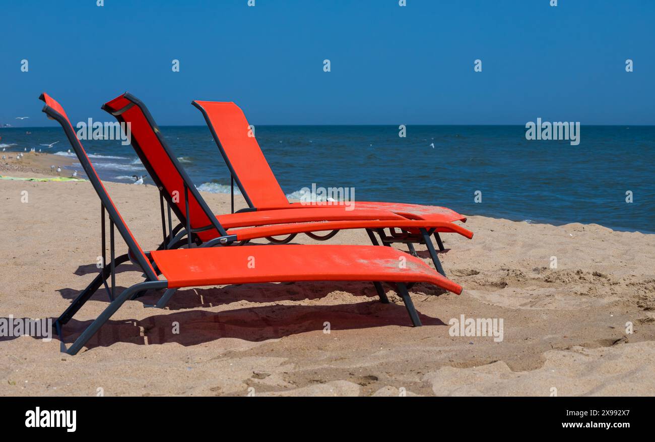 Rote Liegen am Strand. Niemand auf den Liegestühlen. Roter Liegestuhl an einem flachen Sandstrand, Strandausrüstung. Stockfoto