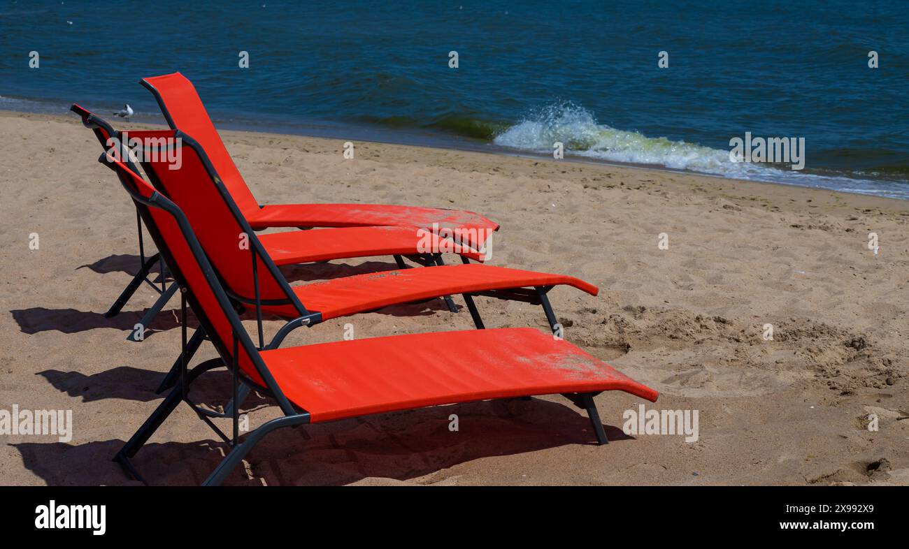 Rote Liegen am Strand. Niemand auf den Liegestühlen. Roter Liegestuhl an einem flachen Sandstrand, Strandausrüstung. Stockfoto