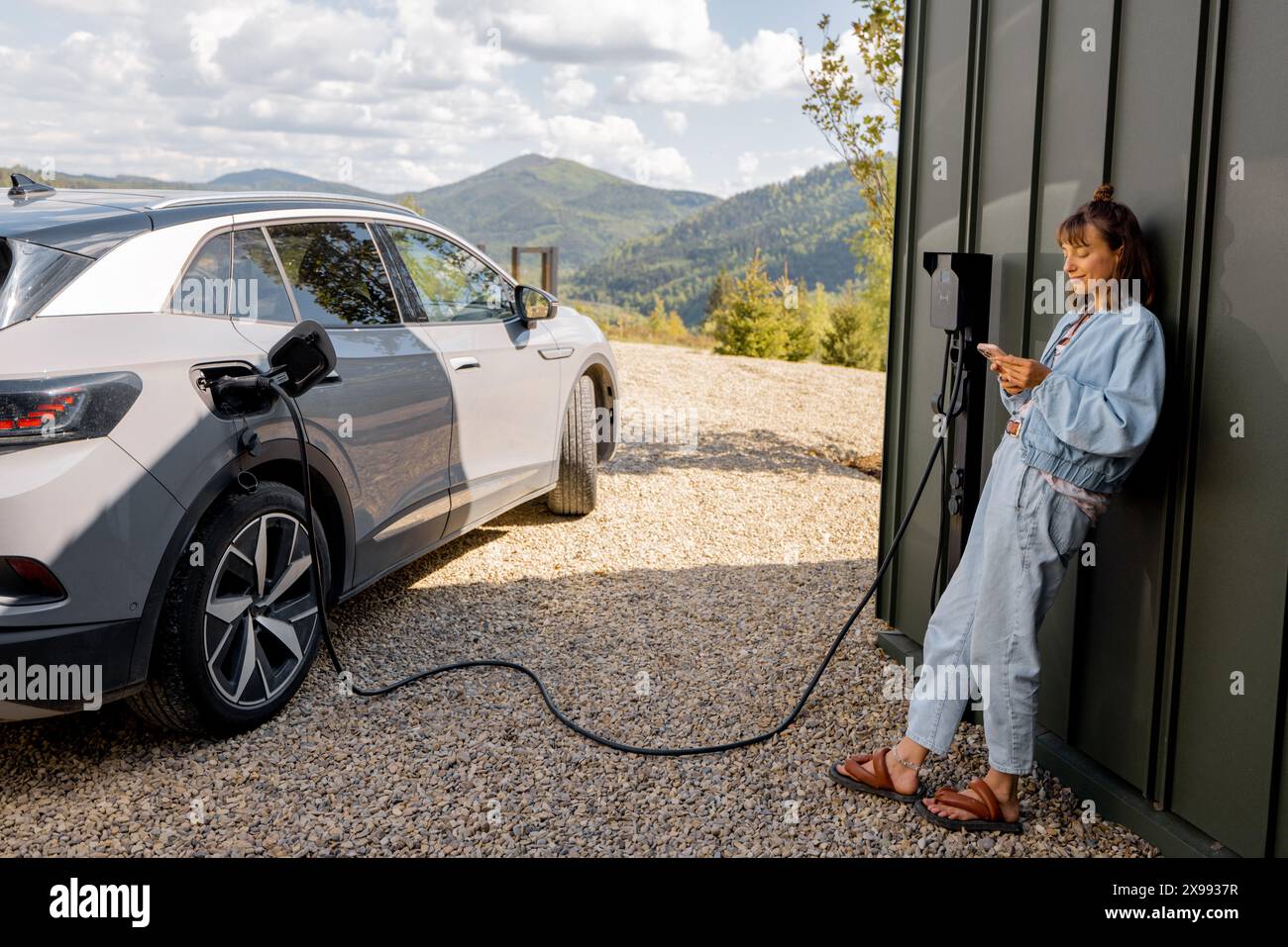 Frau steht mit einem Smartphone und wartet darauf, dass das Elektroauto aufgeladen wird Stockfoto