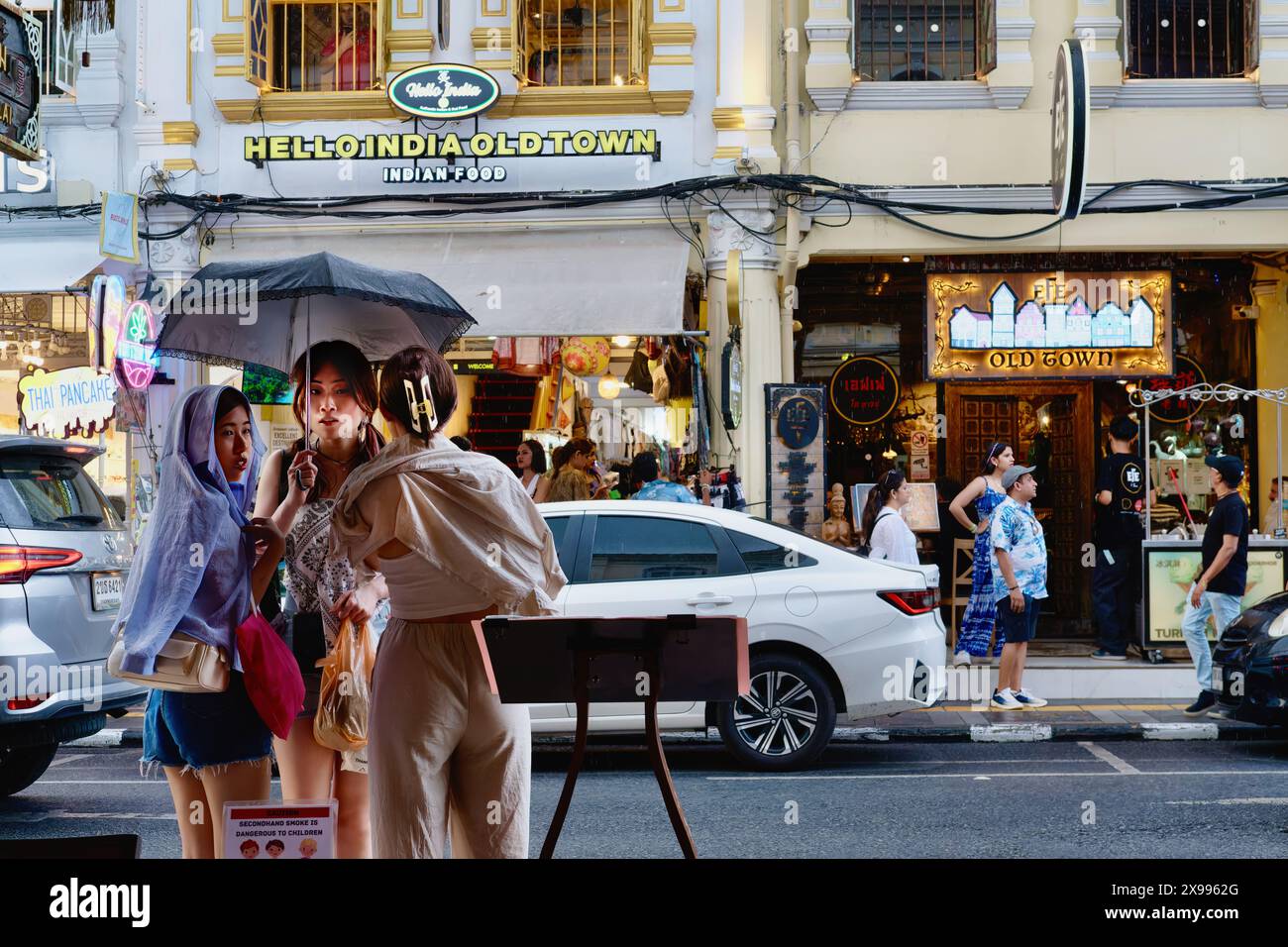 Drei junge thailändische Touristen, die während einer Monsundusche unter einem Regenschirm Zuflucht suchen; in der Touristengegend Thalang Rd, Altstadt, Phuket Town, Thailand Stockfoto